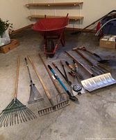 Photo showing red wheelbarrow and various garden hand tools arranged on concrete floor.