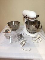 White KitchenAid Ultra Power stand mixer with attached stainless steel bowl, two bowls, whisk, dough hook, flat beater, splash guard, and instruction manual on table.