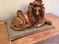 Front angled view of gold painted lion sculpture resting on a green pillow with gold braid, showing detailed mane and paws.
