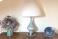 Full frontal view showing the lamp, vase with dried flower arrangement, and ceramic plate on a wooden surface against a wall.