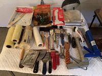 Wide view of painting tools including rollers, brushes, putty knives, caulking guns, scrapers, and a face shield on a quilted cloth on a table