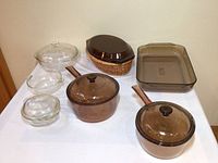 Full set of Pyrex cooking and baking dishes including brown saucepans, clear dishes, and brown oval baking dish with lid and wicker basket stand.