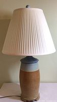 Ceramic table lamp with tall, heavy base in shades of rust and blue, topped with a white pleated lampshade.