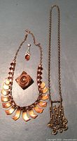 All four jewelry pieces laid out on a surface: copper choker necklace, long copper pendant necklace, copper pin brooch, and sterling silver amber pendant.