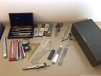 Full view of drafting tools laid flat showing box, compasses, rulers, protractors, lead holders, and small metal tools.