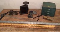 Full lot on wooden table showing antique scale, vise, drill bit sets, metal storage chest, folding ruler, pliers, and sharpening stone.