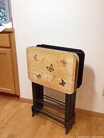 Four vintage folding TV trays with butterfly prints on beige surfaces stacked on black metal stand against wall. Surfaces show wear and scuffing.