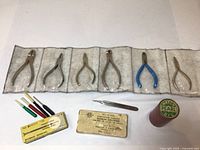 Wide view of six small pliers in clear fabric roll case, five small screwdrivers with colored handles, and a container of Watch Pearl oil on white background.