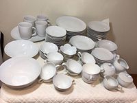 Wide view of the full collection of white dishes including bowls, plates, cups, and serving pieces arranged on a table with a quilted white cover.