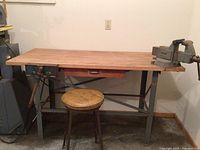 Front view of workbench showing wood top, metal frame, built-in drawer, hold-down clamp on left and bench vise on right