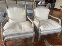 Front view of two off white fabric and light wood living room armchairs with cushions on a carpet.