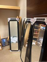 Four black over-the-door mirrors in original packaging with cardboard protectors at corners, staged leaning against a wall in a room.