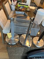 Three different metal paper towel holders on a kitchen counter alongside metal trays and wire baskets.