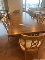 Wide angle photo showing large rectangular wooden dining table with 8 matching chairs in a well-lit room with city view background.