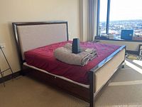 King size bed with dark wood frame and beige fabric panels on headboard and footboard, red sheet on mattress, gray blanket folded on top.