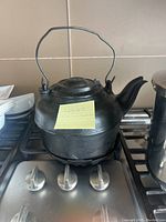 Front and side views of a large cast iron tea kettle with a wire handle and spout placed on a stove burner, showing the kettle details and note on it.