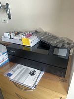 Printer on wooden surface with ink cartridges and manuals visible on top