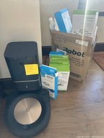 Two rose gold Roomba s9 robots, black Clean Base dock, boxes of replenishment kits, Braava jet pad packs and cleaning solution bottles arranged on floor