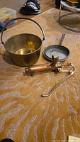 Brass pot with handle, vintage copper skillet, and vintage brass and copper water hand pump arranged on carpet.