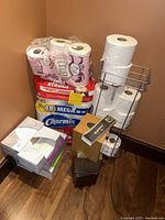 Photo showing stacked packs of toilet paper (Kirkland, Charmin, Angel Soft), Kleenex tissue boxes, a wooden tissue box holder, metal tissue holder rack, and small metal trash can.