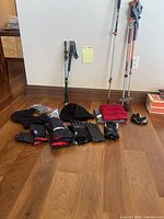 Various men's winter gloves, black winter hats, and face masks laid out on wood floor with walking sticks leaning against wall.