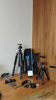 All three tripods arranged on a wooden desk surface along with multiple miscellaneous camera accessories such as mounting plates, clips, and fasteners in black metal and plastic.