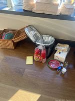Overview of wicker picnic basket with picnic items and Coleman soft-side cooler with various paper goods and picnic supplies on the floor by a window.