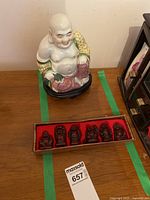Front view of the porcelain Laughing Buddha statue with six small Buddha figurines in their red-lined box in front of it on a table.