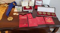Overview of lot showing medals with blue ribbon, four large coins in display box, red envelopes with certificates and coins laid out on table