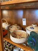 Display cabinet shelf showing multiple covered bowls and teapots with various designs and a greenish ceramic jar.