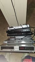 Stack of six video/DVD players: Sony Betamax SL-2401 at bottom, above are various DVD players in black and silver (Philips, Toshiba, Panasonic, Koss). Power cords visible, units look used but untested.