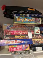 Shelf with stacked game boxes including Crossfire, Hotel, Frog Soccer, Grape Escape, HeroQuest, Battle Masters, Girl Talk, Fashion Doll Salon Play Set, and Wildlife puzzle