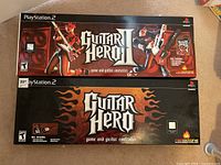 Two Guitar Hero PS2 bundle boxes for Guitar Hero II (top) and Guitar Hero (bottom) on carpet