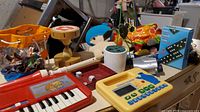 Photo showing an orange and beige wooden Den Den Daiko hand drum on a table, part of a red toy keyboard, dice, a yellow electronic learning toy with buttons, and a blue plush toy, with various other toys scattered.