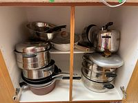 Wide view of cupboard showing multiple stacked pots, pans, lids, and bowls including a large steamer pot on right side.