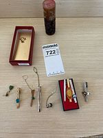 Overview photo showing seven tie clips with different designs laid out on a wooden table including a red presentation box and a red cylinder stamp excluded from sale.