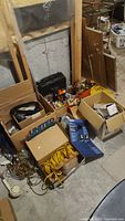 Photo of several cardboard boxes filled with tools, ropes, cords, tapes, and electrical hardware on concrete floor.