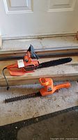 Both Black & Decker electric tools together on a cement doorstep; chainsaw with long blade and hedge trimmer below it