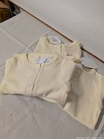 Three cream-colored ribbed fabric baby onesies folded, showing the button placket at the chest and tags for new old stock status.
