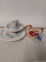 Photo showing a Cabbage Patch Kids ceramic tea cup, saucer, and plate set along with a Cabbage Patch Kids figurine.