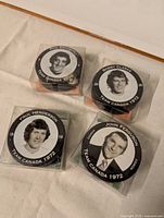 Four collectible hockey pucks with portraits of Phil Esposito, Bobby Clarke, Paul Henderson, and John Ferguson, each in clear display cases on white fabric background.