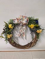 Front view of a fall-themed wreath on a white background featuring a natural grapevine base, mixed greenery, yellow and blue flowers, and a patterned fabric bow at the top.