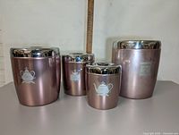 Front view of four metallic pink kitchen canisters with chrome lids showing white decorative silhouettes on three canisters.