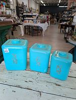 Three teal metal kitchen canisters of different sizes with gold decorative designs on the front, placed on a rustic white wooden surface in a store environment.