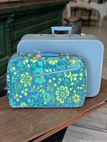 Two vintage luggage pieces: a light blue hard-sided suitcase and a smaller floral-patterned soft-sided suitcase.
