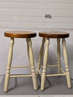 Two wooden stools standing side by side showing round wood tops and cream painted legs with signs of wear.