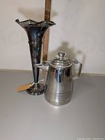 Silver teapot and silver flower vase placed together showing front angles and height contrasted by a ruler in background