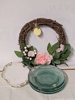 Wicker spring wreath with silk flowers, two green glass plates, and white ceramic dish arranged on a flat surface