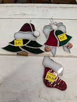 Three stained glass Christmas ornaments displayed on a white surface, each with price tags visible: Santa hat $24, bell $25, stocking $18.50.