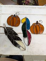 Overhead image showing all four stained glass pieces on a white wooden surface: two orange pumpkins, one colorful parrot, and a dark leaf